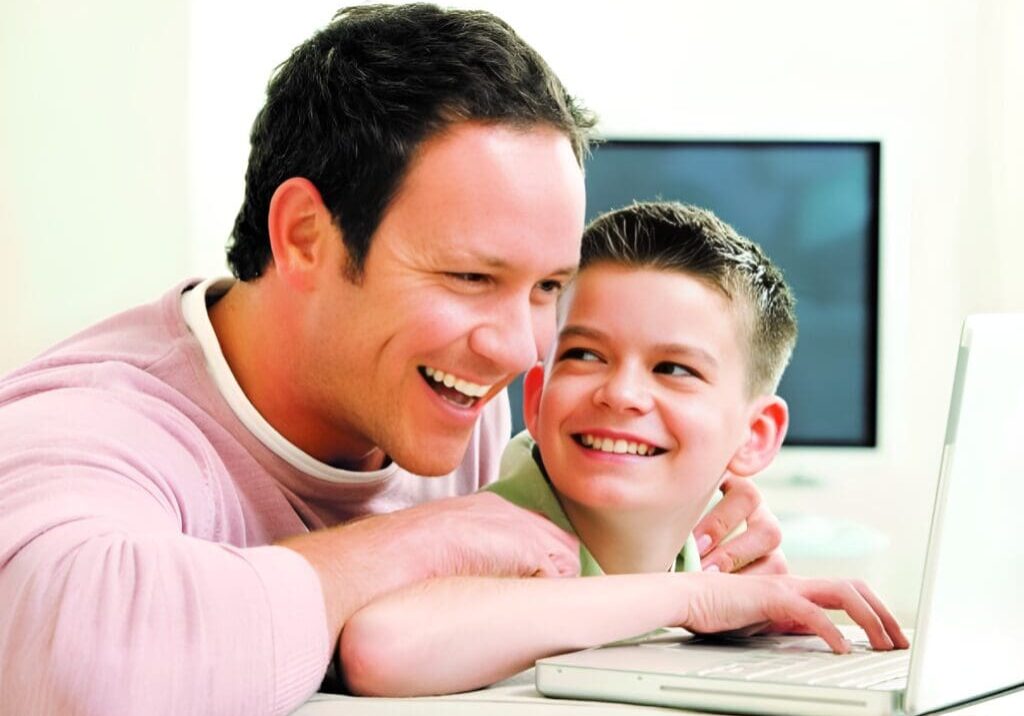 A man and boy smile at the camera while using a computer.